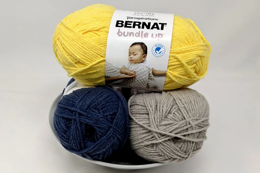 bernat bundle up yarns in duckling, beluga and pigeon stacked together on a white bowl