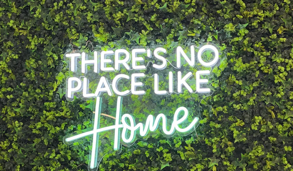 A neon sign says there’s no place like home against greenery 