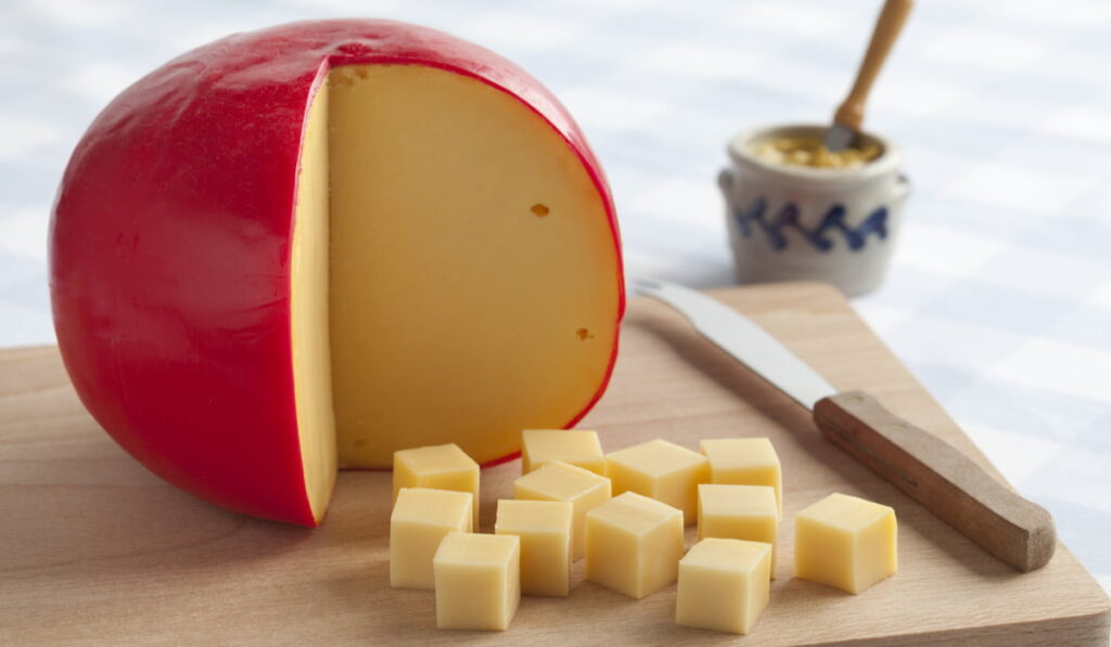 Edam cheese sliced in cube size on a chopping board beside a cheese knife