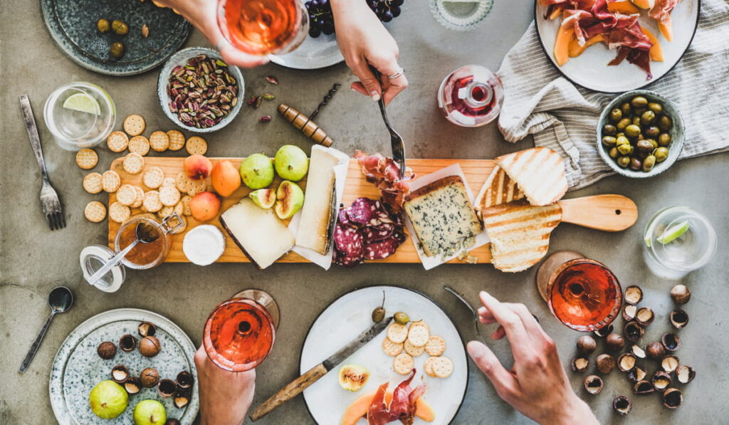 flat lay photo of Flat-lay of charcuterie and cheese board, some nuts, olives, bread and crackers; hand of a male and female holding wineglasses 