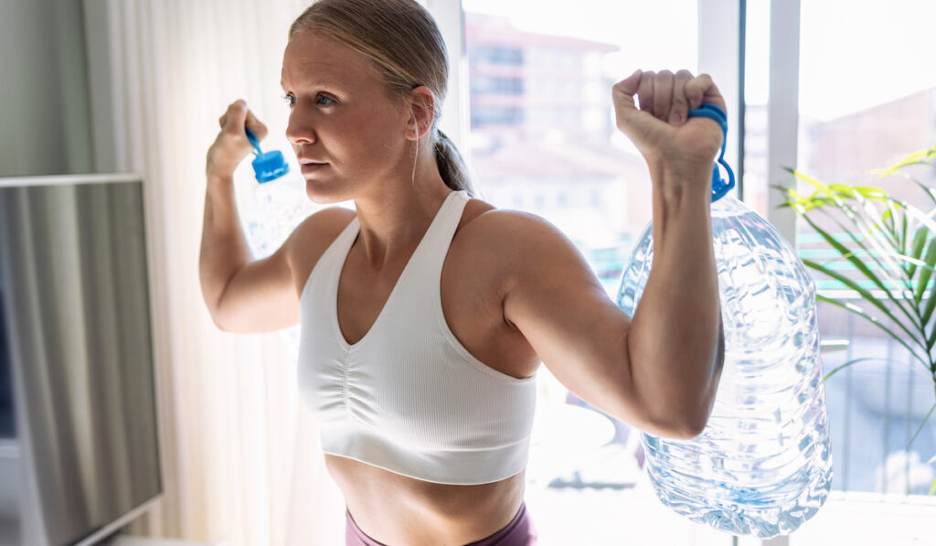 Beautiful gymnast woman doing exercices lifting eight liters water bottles in living room at home 
