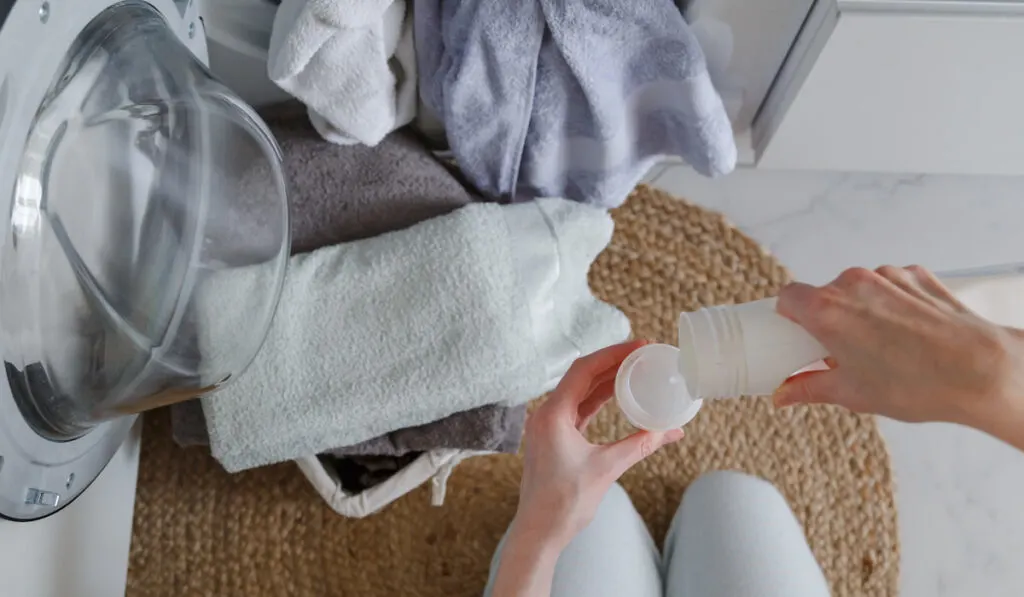 Woman pouring liquid detergent from bottle for laundry in front of washer