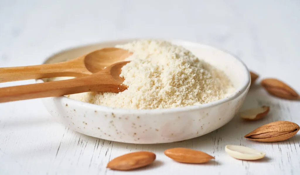 almond flour in white bowl wooden spoon and almonds on white background