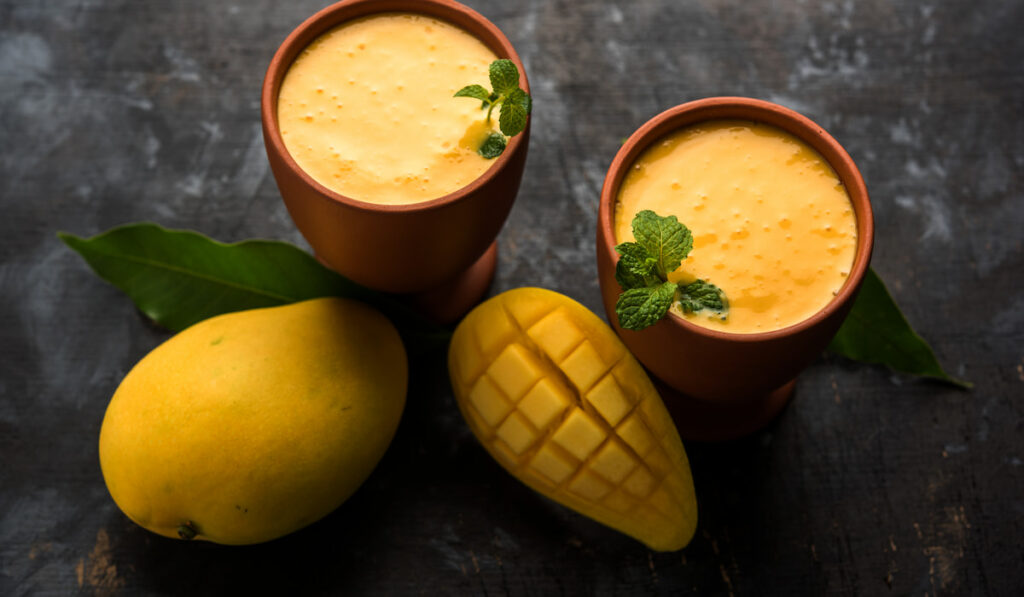 two glass of mango lassi with fresh whole and sliced mango