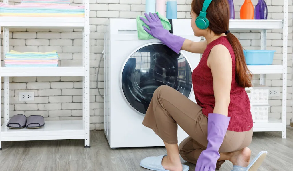 woman listing music by headphone and cleaning washing machine in the laundry room 