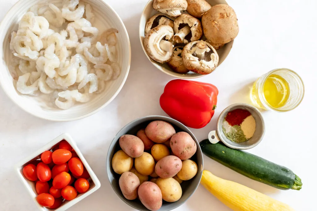 ingredients for grilled shrimp and veggie skewers. bowl of shrimp, mushrooms, potatoes, grape tomatoes, one red bell pepper, two zucchinis, olive oil and some dried herbs on the table
