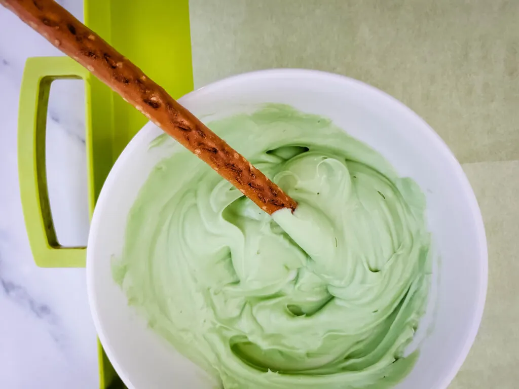 A pretzel rod dip  into the green melted candy in a bowl on green tray