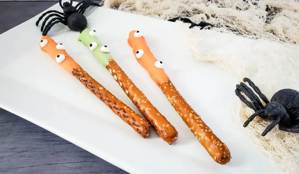 Halloween Monster Pretzels coated with green and orange melted cany on white plate set up and black spiders on the side.