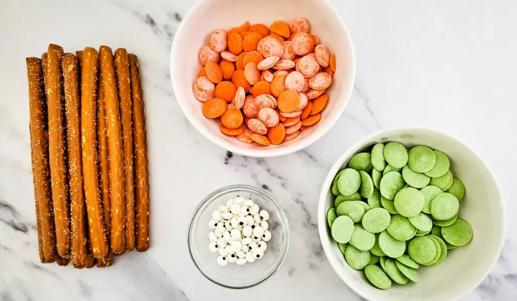 Halloween Monster Pretzels ingredients on a marble table, pretzels, green and orange candy melts and eyeballs in glass bowl