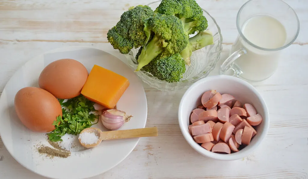 Ingredients of Keto Low Carb Breakfast Casserole, garlic, salt, eggs, cheese, parsley, ground pepper on white plate, sausages, cream and broccoli