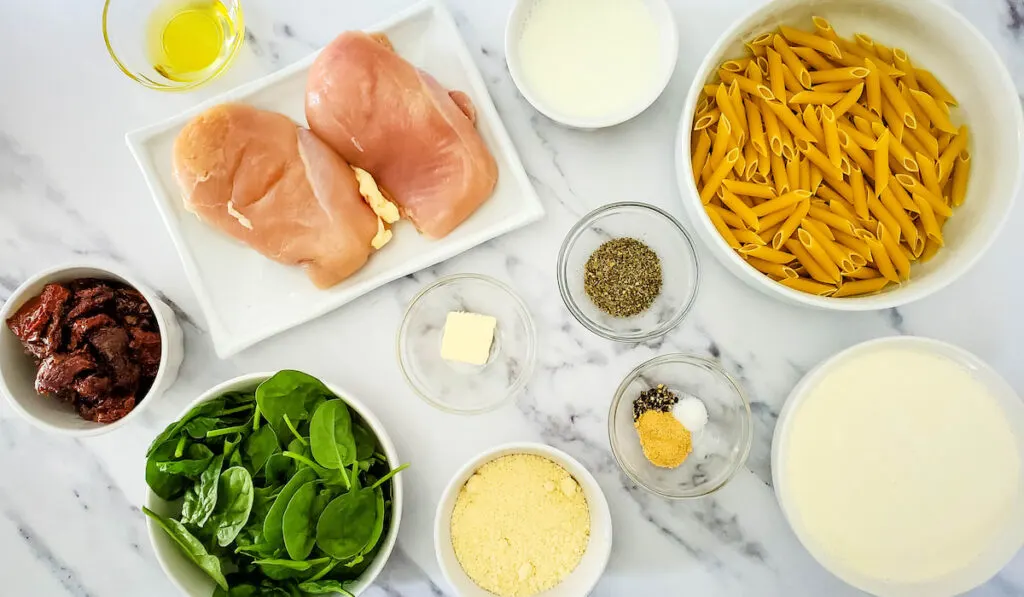 Tuscan Chicken Pasta Ingredients on the table. Penne Pasta, Spinach, chicken breast, oil, butter, salt, pepper, cream, milk all in a bowl
