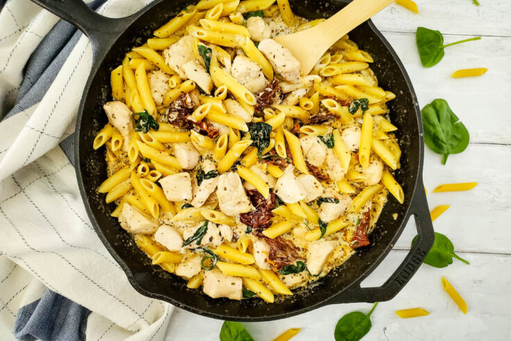newly cooked Tuscan Chicken Pasta in a pan with wooden spoon, along with raw spinach, raw penne pasta and a cloth on a white table