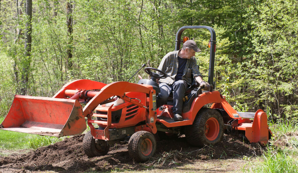 A utility tractor tilling the soil with its operator in the open cab