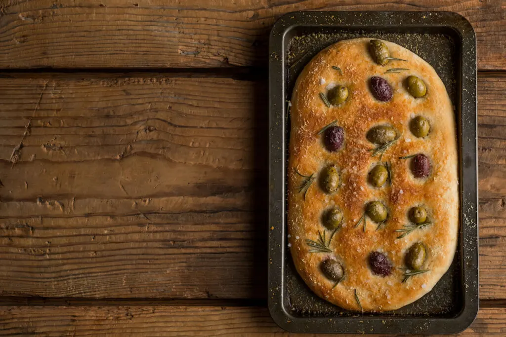 Flat Focaccia Italian bread from dough with olives, oil, parmesan and herbs in a baking roast pan on wooden table