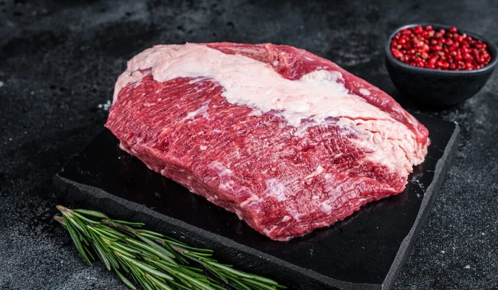 Fresh raw round brisket beef cut n a marble board, black background with spices on the sides