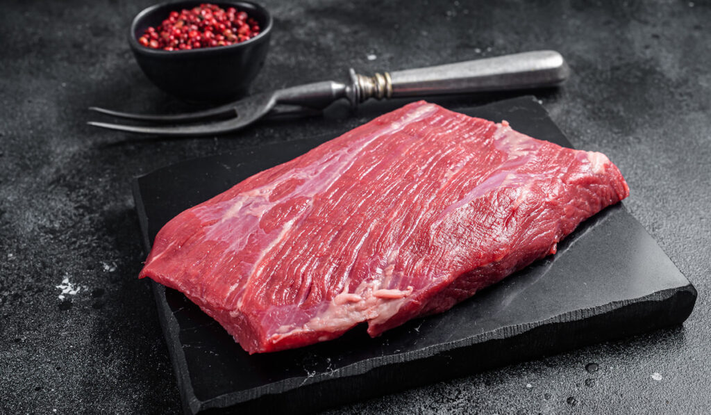 Prime choice flank steak, raw beef meat on marble board with herbs on Black background