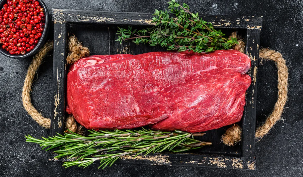 Raw Whole Tenderloin beef meat on a wooden tray with herbs on black stone background