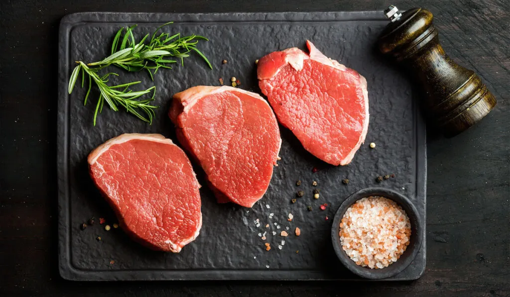 Raw beef Eye Round steaks with spices and rosemary on black slate stone board over dark wooden background