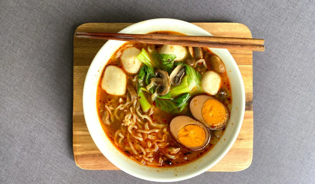 Top view of Japanese ramen spicy soup with fish balls green bak choy and brasied five spice duck eggs in white bowl on serving brown wooden tray
