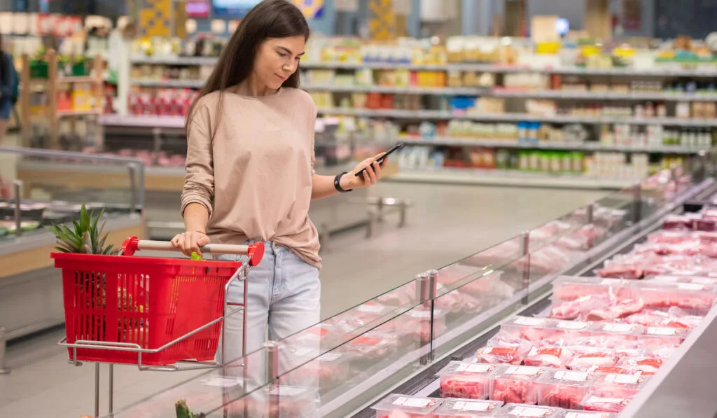 Young woman standing in aisle with shopping cart choosing fresh raw meat in modern supermarket

