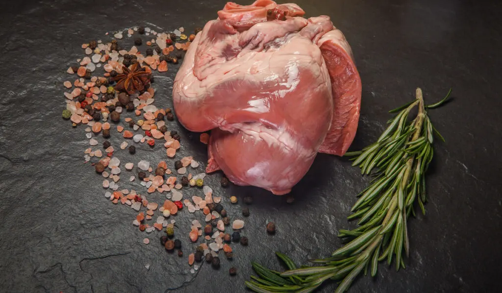 closeup raw beef heart with spices and rosemary on black stone background