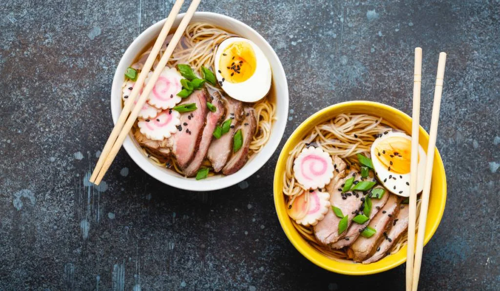 Two bowls of Japanese noodle soup ramen with meat broth, sliced pork, narutomaki, egg with yolk on rustic stone background