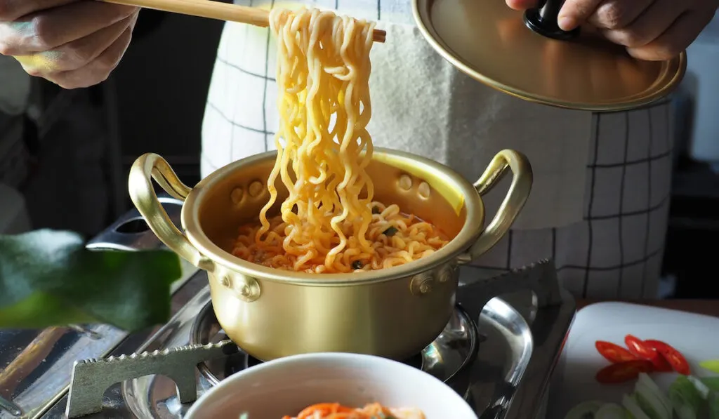 Man cooking ramen in aluminum pot on gas stove over wooden table with kimchi, chili and scallion on the side