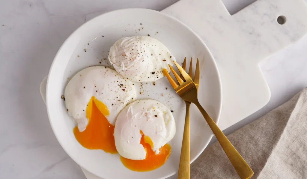 Three poached eggs with egg yolk on a white plate on a marble board and golden forks
