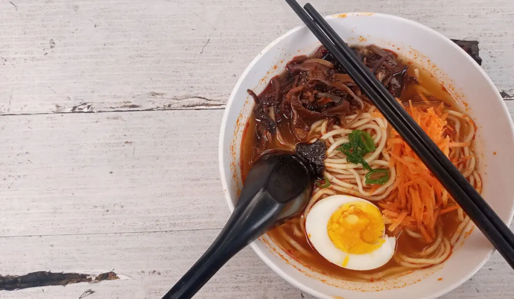 Ramen noodles with boiled egg, grated carrots with a spicy soup, black chopsticks on the table