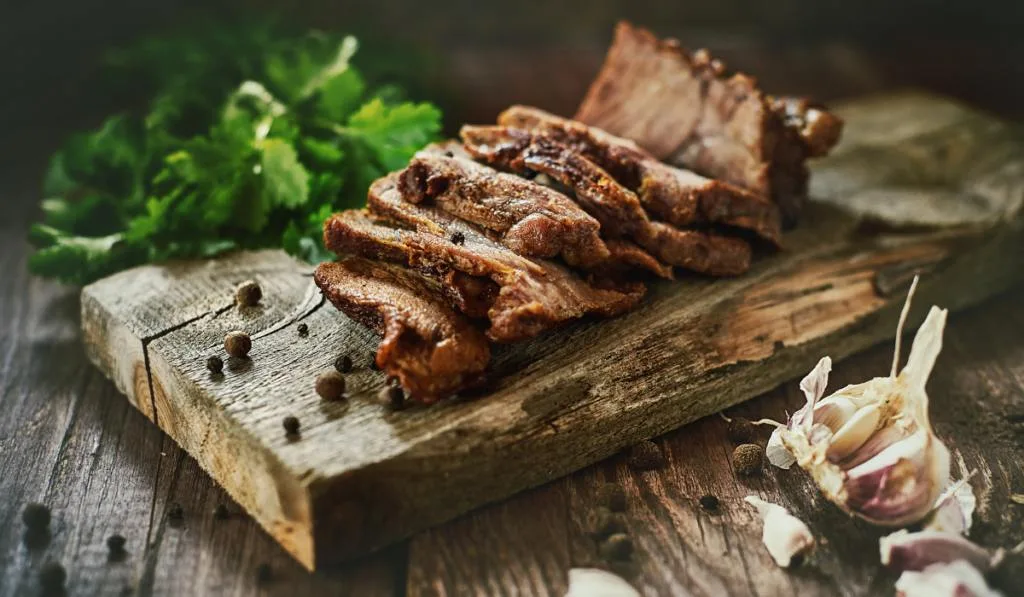 cooked and sliced meat on a wooden chopping board with some spices on the table