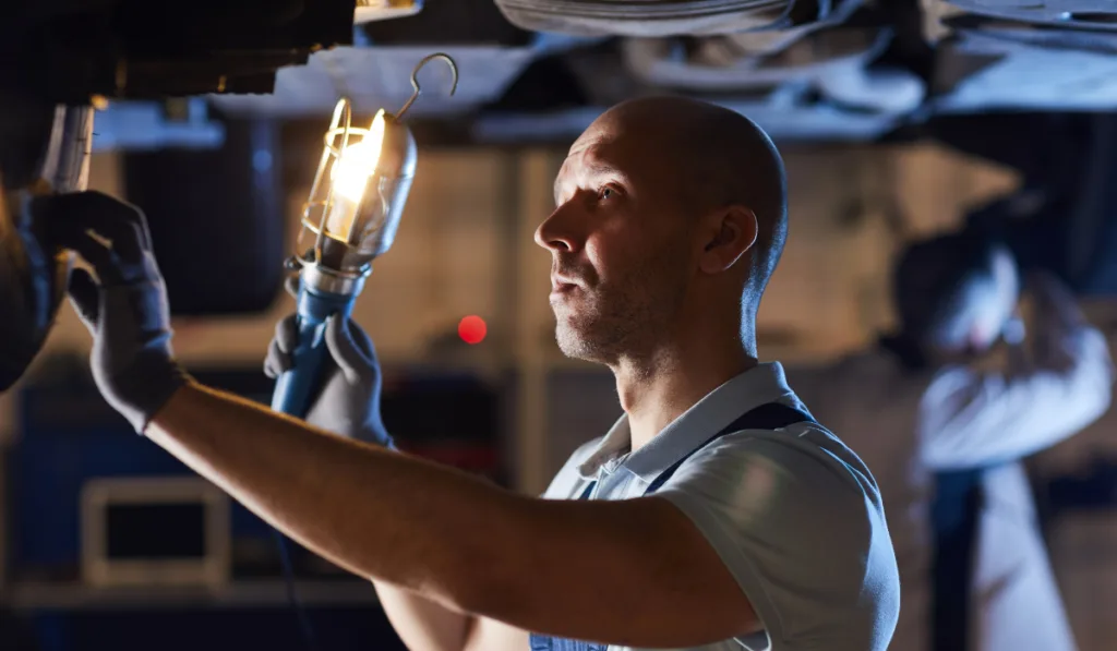 car mechanic inspecting vehicle while holding flashlight lamp under car lift