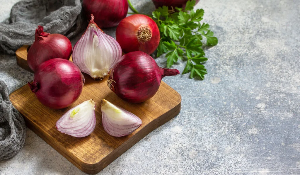 Whole and sliced purple onions on a wooden chop board on a stone countertop