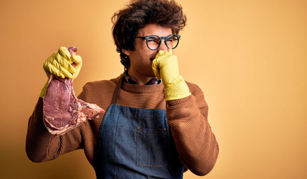 Young butcher man holding stinky meet steak holding breath with fingers on nose on yellow background