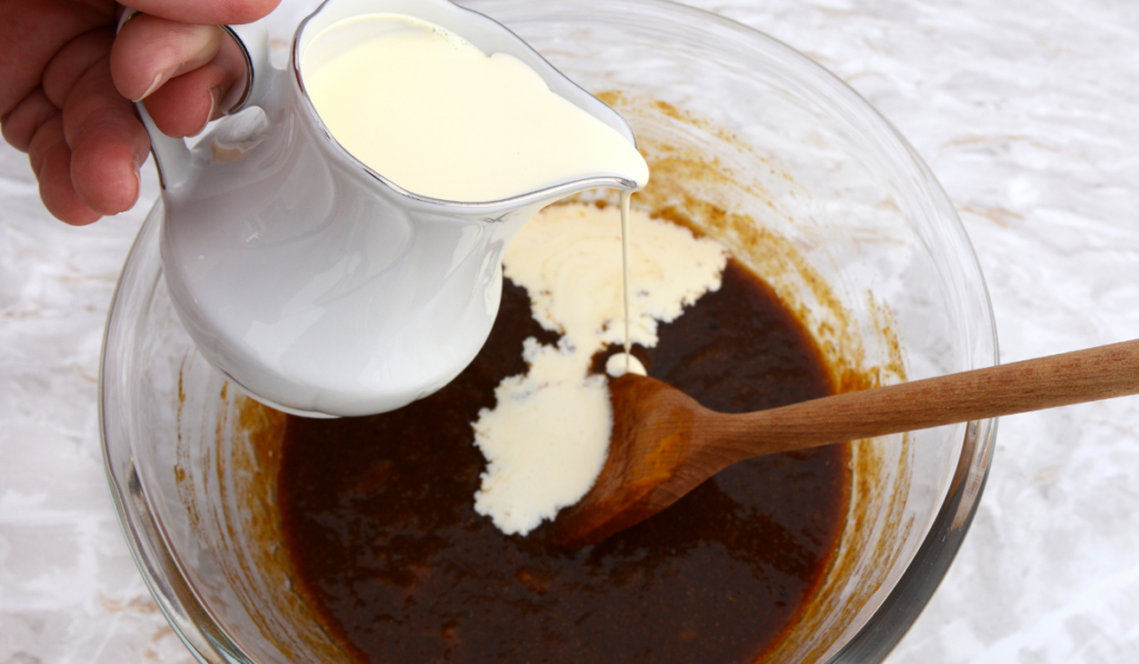 Woman pouring heavy cream from a jug into pumpkin pie filling in a bowl

