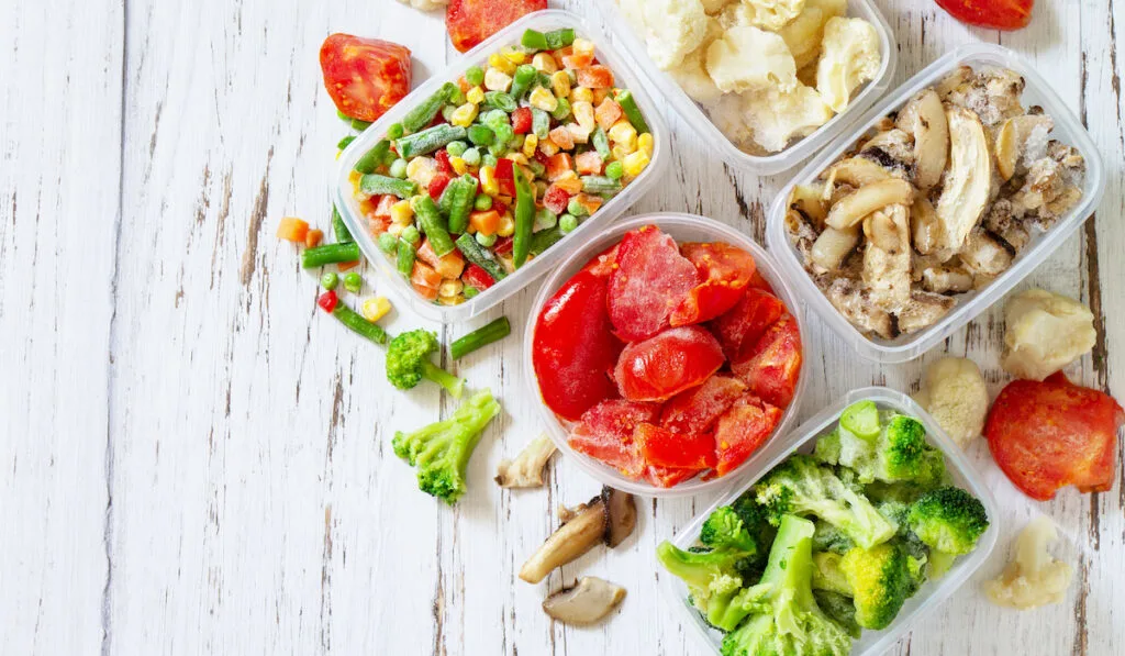 Container with frozen different vegetables and mushrooms on white wooden background