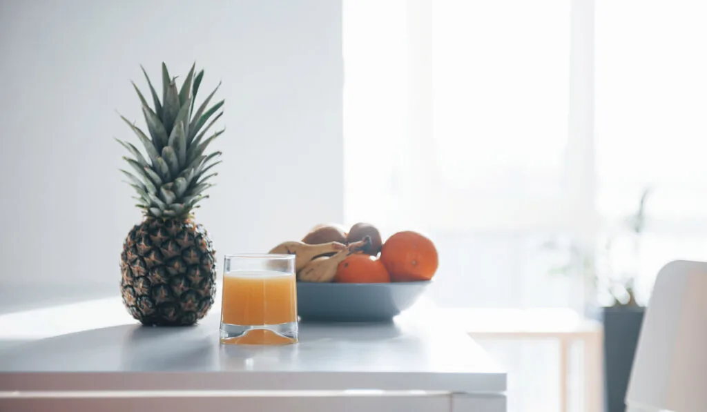Pineapple, bananas, oranges and glass with fresh juice on kitchen countertop