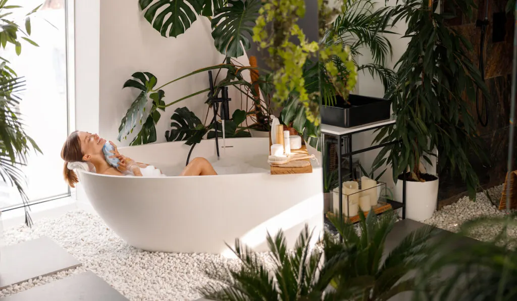 Relaxed young woman bathing in modern bathroom interior decorated with tropical plants
