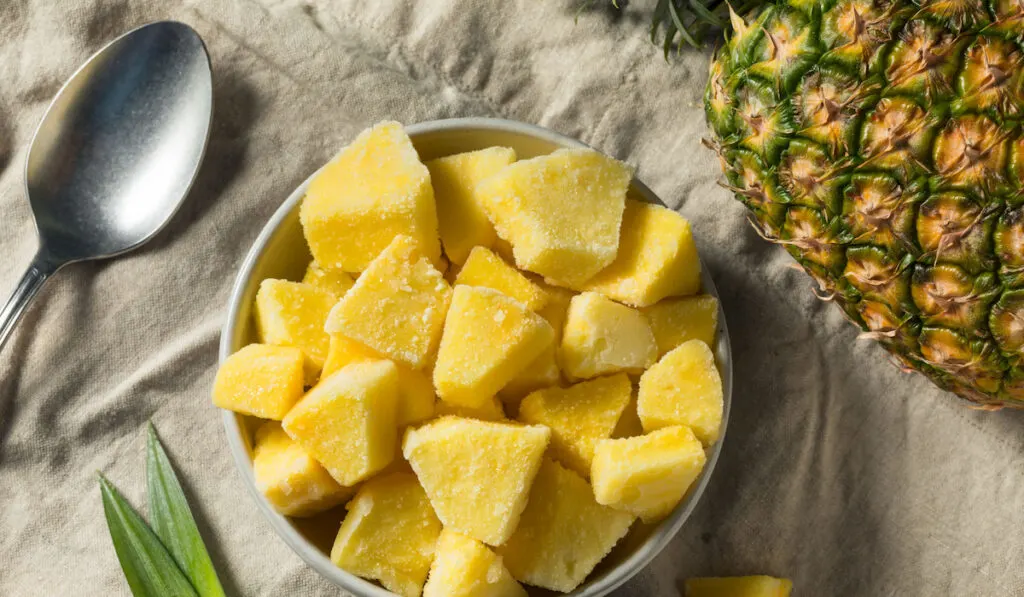 Yellow Organic Frozen Pineapple Slices in a bowl with spoon and whole pineapple on the table
