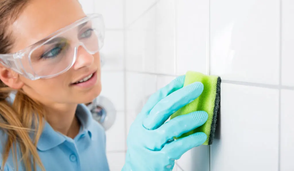 woman with protective eye wear cleaning the white tile of the wall using sponge