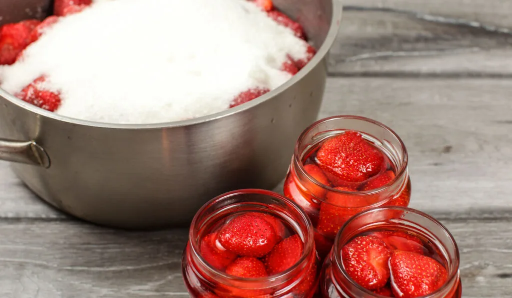 Bottles with homemade pickled strawberries, steel pot with more berries covered with sugar in background