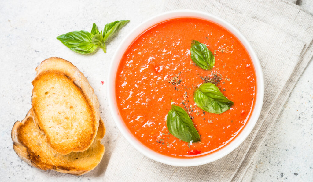 Tomato soup with basil in white bowl with bread on the table